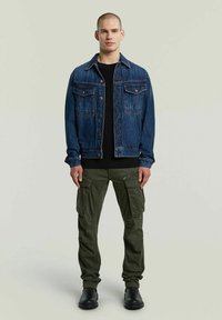 Giacca di jeans di colore blu scuro con due tasche sul petto, abbinata a pantaloni cargo verde oliva con numerose tasche e dettagli con zip. Stivali neri.