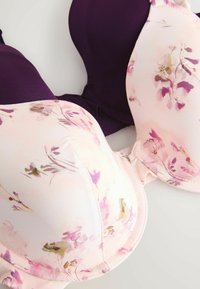 Du liemenėlės pasižymi viena violetine medžiaga, kita – šviesiai rožine su gėlių raštais, demonstruojančiomis lygius paviršius ir struktūruotas formas.