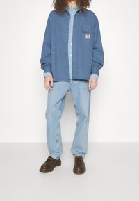 Blauw langmouwen shirt met knoopsluiting aan de voorkant, borstzak, gelaagd over een lichtblauw spijkerhemd; gecombineerd met lichtblauwe jeans en bruine schoenen.