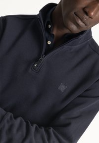 Sudadera marina con cremallera de un cuarto, textura acanalada y parche de logo. Presenta un cuello alzado y una capa interior en contraste.