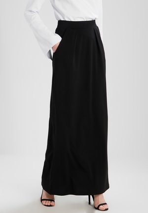 Femme portant une longue jupe noire avec des poches latérales, associée à des sandales à talons hauts noires et une chemise blanche à manches longues avec des poignets évasés.