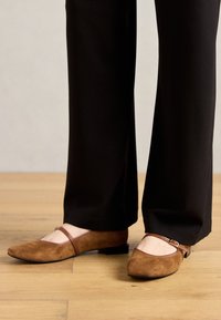 Des ballerines en suède marron avec un bout pointu, une bride Mary Jane et un petit talon, associées à un pantalon noir à jambes larges sur un parquet.