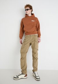 Bruine hoodie met buidel en logo, kaki cargobroek met meerdere zakken, en zwart-witte sneakers met bruine accenten.