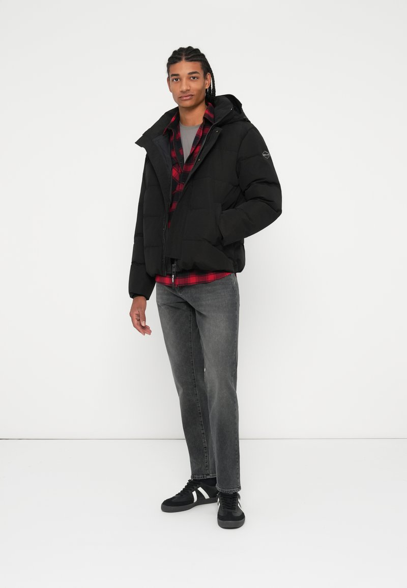 Schwarze Daunenjacke mit Reißverschluss vorne, abnehmbarer Kapuze und gestepptem Design. Getragen über einem rot-schwarz karierten Hemd, kombiniert mit grauen Jeans und schwarzen Turnschuhen.