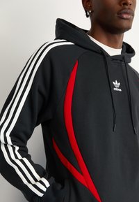 Felpa nera con design a tre righe bianche sulle maniche e dettagli accentuati in rosso. Presenta un cappuccio con cordino e una tasca a marsupio frontale.