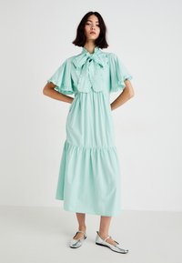 Un vestido midi de color verde menta, con capas y un escote con lazo, y mangas con volantes, hecho de una tela ligera. Unos zapatos planos plateados complementan el atuendo.