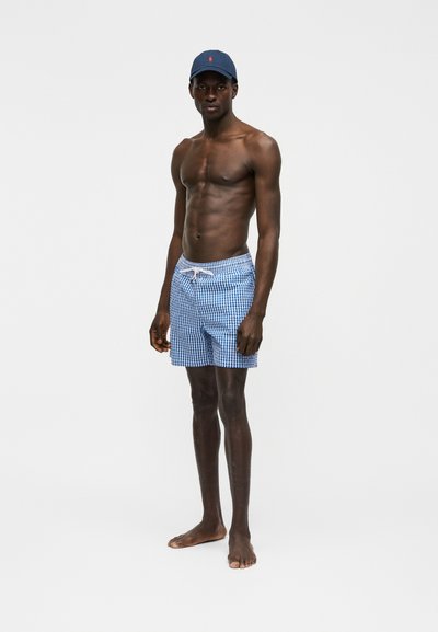 Άνδρας χωρίς πουκάμισο, όρθιος ξυπόλητος, φοράει μπλε και λευκά καρό μαγιό και μπλε σκούφο μπέιζμπολ, σε απλό φόντο.