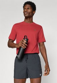 Κόκκινο κοντομάνικο μπλουζάκι με μικρή κεντημένη σχέδιο, σε συνδυασμό με σκούρο γκρι σορτς που διαθέτουν πλαϊνές τσέπες. Μαύρη μπουκάλι νερού με καπάκι.