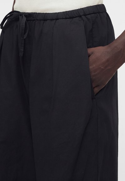 Άτομο που φορά μαύρο παντελόνι με κορδόνι και το χέρι ακουμπισμένο στην πλαϊνή τσέπη, συνδυασμένο με ένα ανοιχτόχρωμο ριμπ τοπ.