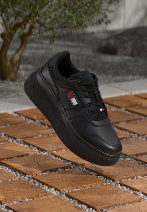 Sneaker casual nera con suola spessa e logo Tommy Jeans, posizionata sopra piastrelle di legno con fessure di ghiaia all'aperto vicino a un albero.