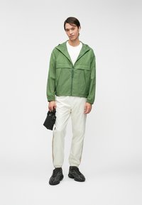 Ελαφρύ πράσινο μπουφάν με φερμουάρ και κουκούλα, με μπροστινή τσέπη. Συνδυάζεται με λευκά φόρμα που έχουν καφέ πλαϊνές ρίγες και μαύρα αθλητικά παπούτσια.