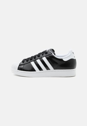Baskets Adidas noir et blanc avec lacets noirs, trois bandes blanches sur le côté, semelle en caoutchouc blanche et design à bout coquille.