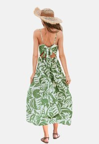 Vestido maxi con un estampado floral verde, que presenta tirantes finos, un lazo en la parte trasera y una silueta fluida. Llevado con sandalias planas y un sombrero de paja.