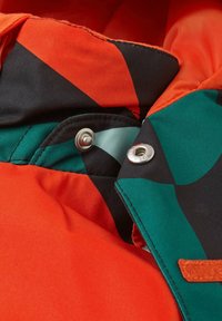Giacca colorata con tessuto arancione, nero e teal, caratterizzata da una texture liscia. Include bottoni decorativi e una fodera a motivo.