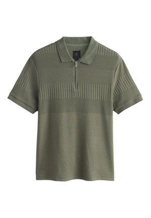 Mann in olivgrünem Poloshirt und hellgrauer Hose lehnt an einer beigen Wand, hält eine schwarze Tragetasche und steht neben einer großen Topfpflanze.