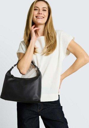 Lachende vrouw met lang blond haar draagt een wit kortärmelig shirt en donkere broek, houdt een grote zwarte leren handtas op haar schouder.