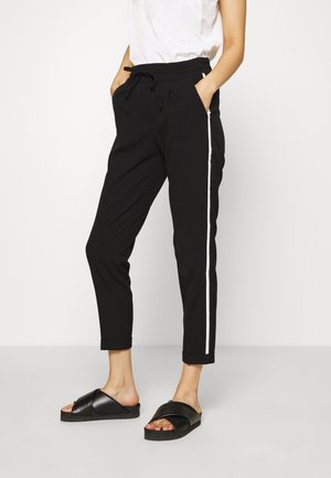 Pantalons noirs à taille élastique avec poches latérales, arborant une bande blanche le long de chaque jambe. Portés avec des sandales plates noires.