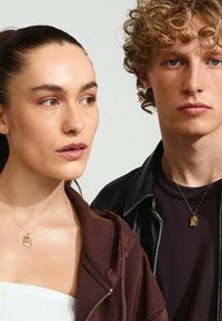 Zlaté náhrdelníky s príveskami na dvoch modeloch. Jeden má na sebe hnedú zipsovú mikinu; druhý má čiernu koženú bundu cez tmavé tričko.