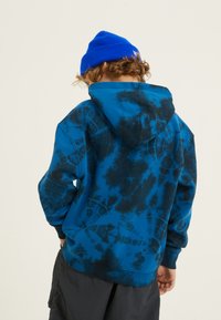 Felpa con cappuccio blu tie-dye caratterizzata da motivi neri, un cappuccio ampio e polsini a costine. Un berretto lavorato a maglia blu completa l'outfit.