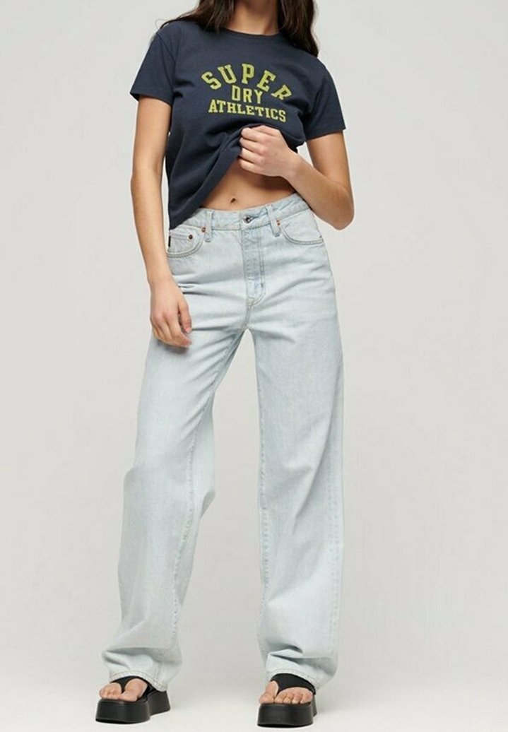 Jasnoniebieskie dżinsy o szerokim kroju, zestawione z granatową koszulą z żółtym napisem. Stylizacja obejmuje czarne sandały na platformie.