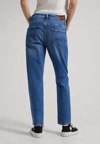 Blå denimjeans med en avslappnad passform, med två bakfickor, en brun läderlapp och synliga sömnadsdetaljer.