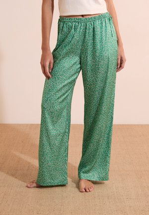 Femme portant un pantalon ample vert à fleurs et un haut blanc, debout pieds nus sur un sol tissé beige.