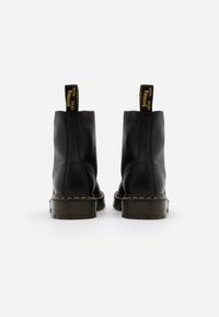Paire de bottines en cuir noir avec semelles en caoutchouc épaisses et coutures jaunes, vues de dos sur un fond blanc.