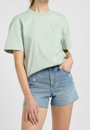 Kobieta ubrana w jasnozieloną, oversize koszulkę i wysokie jeansowe szorty z małym, białym haftem kwiatowym na kieszeniach.