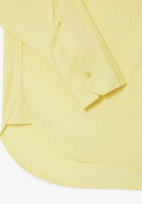 Poignet de chemise jaune à manches longues avec bouton, montrant la couture de la manche et l'ourlet courbé de la chemise sur une surface plane.