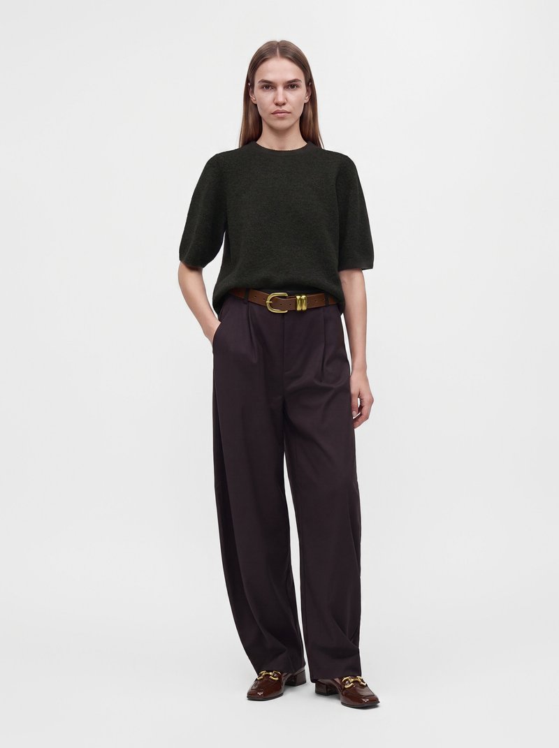 Femme debout portant un pull vert foncé à manches courtes, un pantalon large noir avec une ceinture marron et des mocassins marron avec des détails dorés.