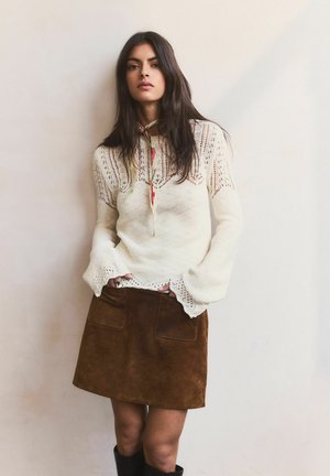 Junge Frau mit langen dunklen Haaren, die einen cremefarbenen Strickpullover, einen braunen Wildlederrock und einen dünnen gemusterten Halstuch trägt und vor einer schlichten Wand steht.