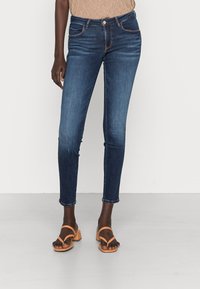 Jeans ajustados de color azul oscuro con una textura suave, diseño de cinco bolsillos y costuras en contraste de color naranja. Con bajos enrollados y combinados con sandalias de tacón bloque en color marrón claro.