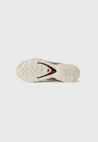 Semelle de chaussure présentant une surface en caoutchouc texturé, principalement blanc cassé avec un accent noir et un détail triangulaire rouge.