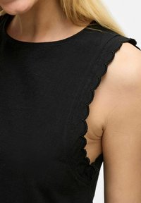 Haut sans manches noir avec un bord festonné sur l'épaule, fabriqué en tissu texturé, présentant un col rond et une coupe ajustée.