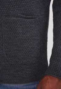 Pullover in tessuto grigio scuro con trama e tasca anteriore. Presenta maniche lunghe e dettagli a costine sui polsi. Stile casual.