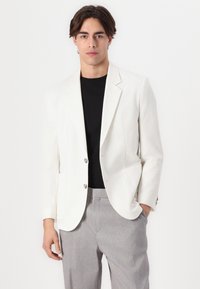 Weißer strukturierter Blazer mit zwei silbernen Knöpfen, kombiniert mit einem schwarzen Hemd und hellgrauen Hosen; hat ein strukturiertes Design und eine schmale Passform.