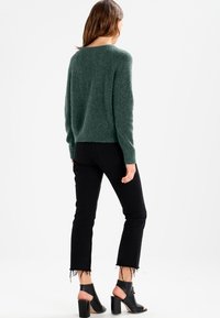 Pull vert à texture duveteuse avec une coupe décontractée, associé à un jean noir évasé avec des ourlets bruts et des sandales à talons noires.