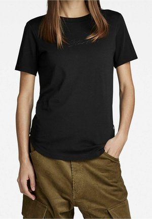 Frau, die ein schlichtes schwarzes kurzärmeliges T-Shirt und braune Cordhosen trägt, mit einer Hand in der Tasche und glatten Haaren.