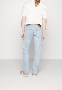 Jeans de mezclilla azul claro con corte recto, que cuentan con bolsillos traseros, una etiqueta de cuero en la cintura y se llevan con una camiseta beige y zapatillas color tostado.