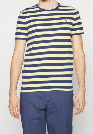 Persona con una camiseta de rayas horizontales amarillas y azul marino de manga corta y pantalones azul marino, de pie con los brazos relajados a los lados.