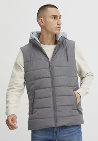 Grå polstret vest med glidelås foran og hette, med sidelommer og quiltet design. Båret over en lysegrå genser med rund hals.