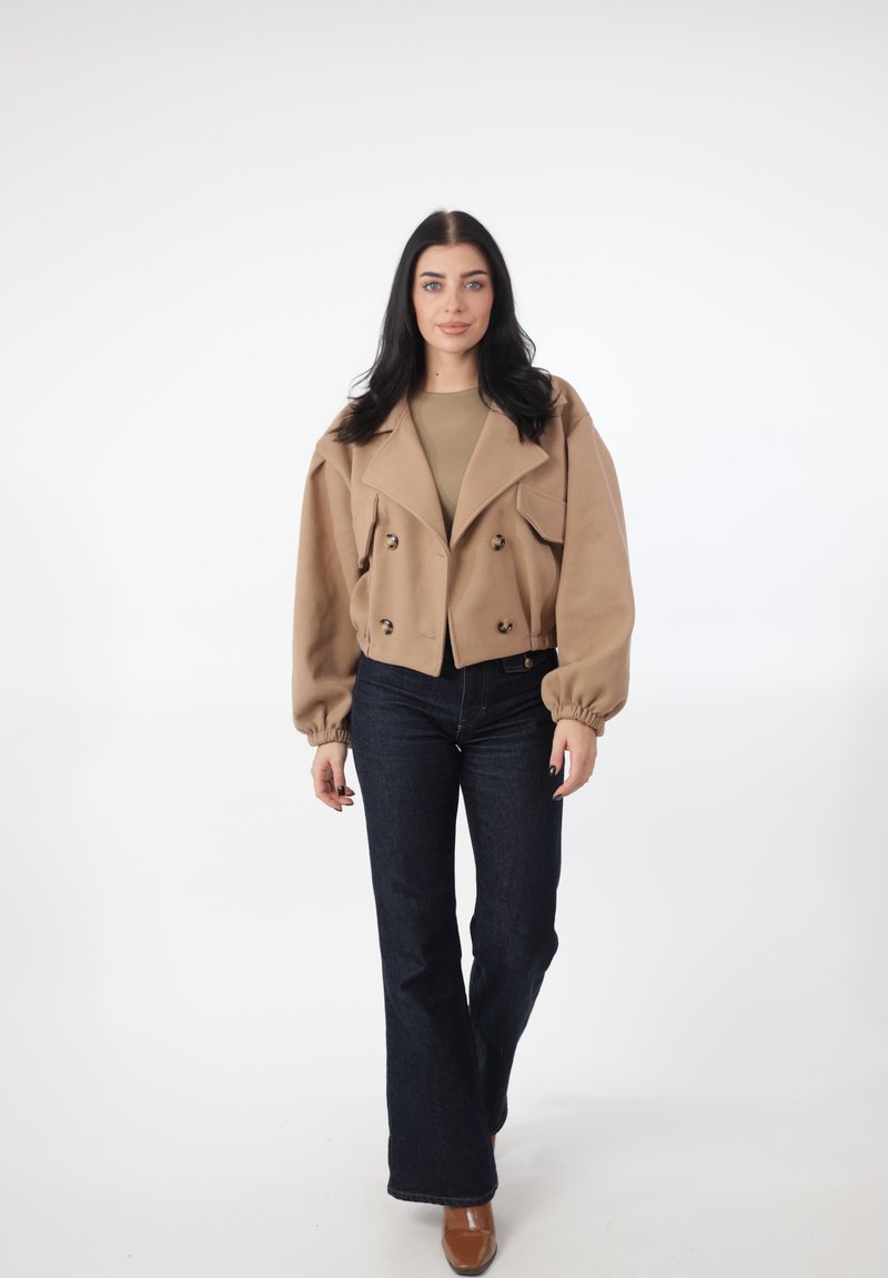 Beiger Wollblazer mit großen vorderen Knöpfen, breiten Revers und aufgepufften Ärmeln; kombiniert mit dunklen, gerade geschnittenen Jeans und braunen Absätzen.