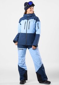 Niebieska i granatowa kurtka narciarska z wysokim kołnierzem, zapinanymi na zamek kieszeniami oraz dopasowanymi spodniami z czarnymi akcentami i luźnym krojem.