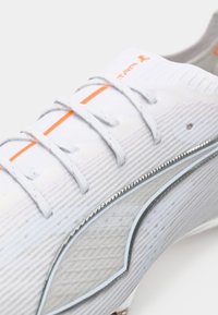 Baskets blanches avec une tige texturée, accents gris, détails orange sur les lacets et un logo sur le côté. Présente un design slim et élancé.