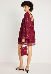 Μπορντό διαφανές φόρεμα με λουλουδάτη κεντητική, μακριά μανίκια και ανοιχτή πλάτη. Συνδυασμένο με μια κόκκινη τσάντα κεντημένη με παγιέτες και μαύρες ψηλές σαμπάλ. 