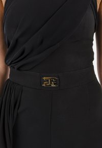 Abito nero con corpetto drappeggiato e una fibbia geometrica in tonalità oro in vita. Presenta un tessuto liscio e pieghe strutturate.