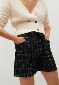 Žena oblečená v krémovém dvouřadovém svetru se zlatými knoflíky a černé kostkované sukně z tkaniny s vysokým pasem, kapsami a třásněmi na lemu.