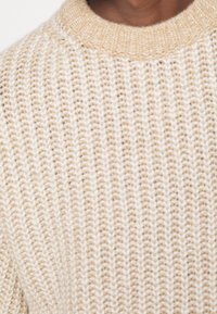 Nahaufnahme einer Person, die einen cremefarbenen und beigen Rippenstrickpullover mit Rundhalsausschnitt trägt.