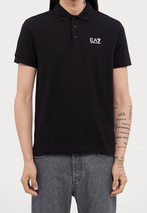 Person trägt ein schwarzes EA7 Emporio Armani Poloshirt und graue Jeans, zeigt Tattoos auf dem rechten Unterarm.