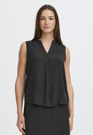 Žena s rovnými hnedými vlasmi, oblečená v čiernej bezrukávovej blúzke s výstrihom do V a čiernej sukni, stojaca pred jednoduchým bielym pozadím.